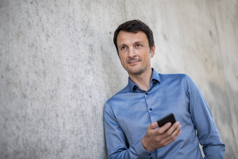 Portrait of smiling businessman with cell phone leaning against concrete wall stock photo