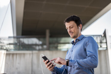 Smiling businessman with cell phone and earphones outdoors - DIGF04713