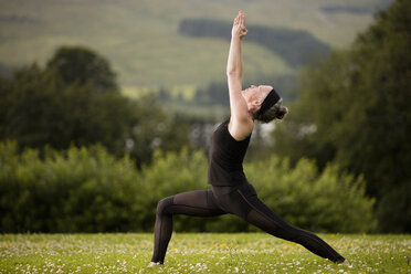 Mature woman practicing yoga warrior pose with arms raised in field - CUF32627