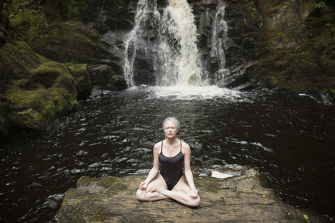 Ältere Frau übt Lotus-Pose vor einem Wasserfall, lizenzfreies Stockfoto