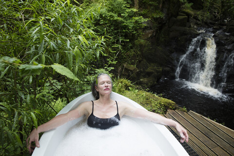 Mature woman relaxing in bubble bath in front of waterfall at eco retreat stock photo