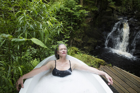 Reife Frau entspannt sich im Schaumbad vor einem Wasserfall im Öko-Retreat, lizenzfreies Stockfoto