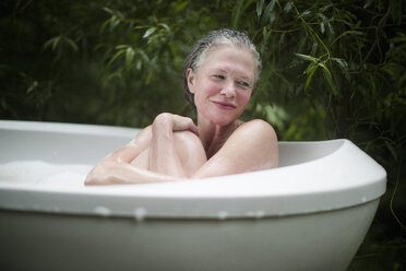 Mature woman hugging knees in garden bubble bath at eco retreat - CUF32668