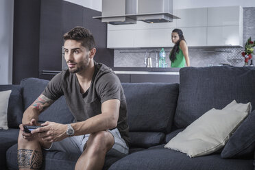 Junger Mann auf Sofa mit Spielkonsole, beobachtet von wütender Freundin - CUF32960