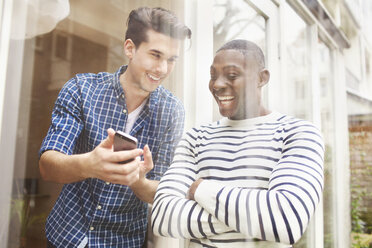 Two young male friends looking at smartphone behind patio glass - CUF33057