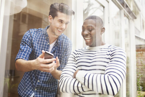 Zwei junge männliche Freunde schauen auf ihr Smartphone hinter einer Glasscheibe im Innenhof - CUF33057