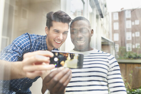 Zwei junge männliche Freunde machen ein Selfie hinter Glas im Innenhof - CUF33059