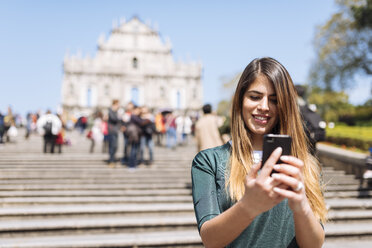 Young female tourist reading smartphone texts in front of St Pauls Cathedral ruins, Macau, Hong Kong, China - CUF33157