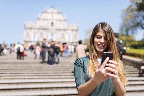 Junge Touristin, die vor den Ruinen der St.-Pauls-Kathedrale auf ihrem Smartphone Texte liest, Macau, Hongkong, China - CUF33157