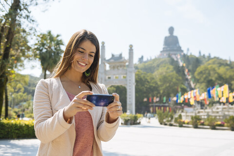 Junge Touristin beim Betrachten von Smartphone-Fotos vor dem Tian Tan-Buddha, Po Lin-Kloster, Lantau Island, Hongkong, China, lizenzfreies Stockfoto