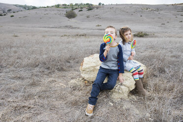 Portrait of brother and sister sitting on rock licking lollipops - ISF10620