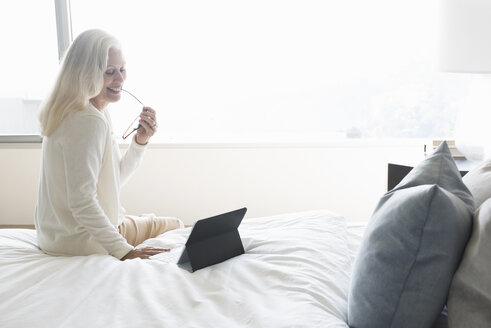 Ältere Frau sitzt auf dem Bett und schaut auf einen Tablet-Computer - ISF10632