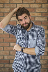 Portrait of bearded young man in front of brick wall scratching his head - AWF00059