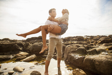 Junger Mann trägt seine Freundin über ein Felsbecken am Strand - ISF10712