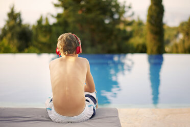 Rear view of teenage boy wearing headphones sitting at infinity pool - ISF10949