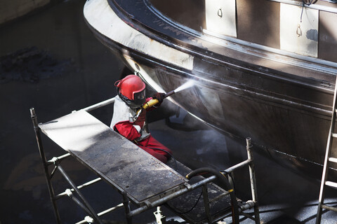 Arbeiter reinigt den Rumpf eines Bootes mit einem Hochdruckschlauch in einer Werft, lizenzfreies Stockfoto