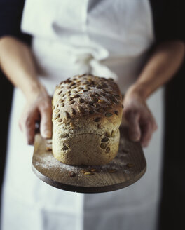 Hände einer Bäckerin, die ofenfrisches Bio-Brot mit Samen auf einem Brett hält - ISF11167