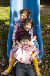 Geschwister in einer Reihe zwischen den Beinen auf der Spielplatzrutsche lächelnd - ISF11226