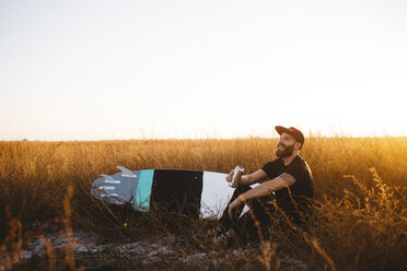 Männlicher Surfer trinkt Bier in einem Feld mit langem Gras bei Sonnenuntergang, San Luis Obispo, Kalifornien, USA - ISF11659