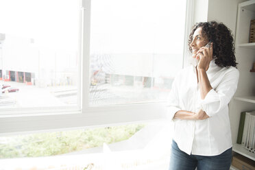 Mature woman talking on smartphone at apartment window - ISF11774