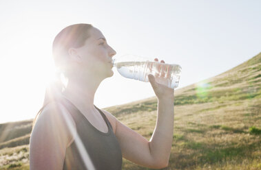 Young female runner drinking bottled water on sunlit hillside - ISF11859