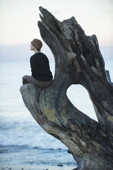 Frau sitzt und schaut von einem großen Treibholz-Baumstamm am Strand aus - ISF11939
