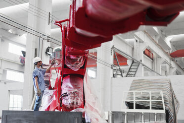 Männlicher Fabrikarbeiter sprüht einen Kranarm in einer Fabrikhalle rot an, China - ISF12092