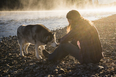 Frau sitzt am Ufer des Bitterroot River und streichelt ihren Hund, Missoula, Montana, USA - ISF12130