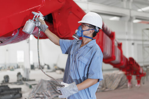 Männlicher Arbeiter sprüht einen Kranarm in einer Fabrikhalle rot an, China - ISF12212