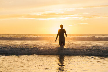 Rear view of mid adult nude womans silhouette walking into ocean at sunset - ISF12355