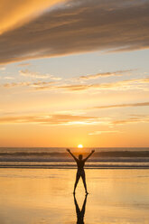 Rear view of mid adult nude womans silhouette standing on beach at sunset, legs apart, arms raised - ISF12356