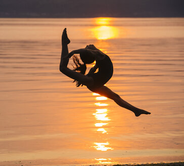 Seitenansicht eines Mädchens in Silhouette am Meer bei Sonnenuntergang in der Luft, die Beine gespreizt, den Kopf zurückwerfend - ISF12358