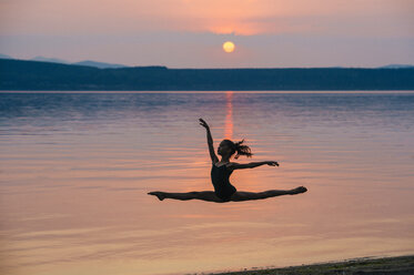 Side view of girl by ocean at sunset leaping in mid air, arms raised doing the splits - ISF12360