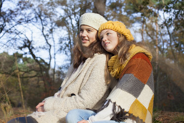 Teenage girls wearing knitwear sitting side by side in forest looking away smiling - ISF12548