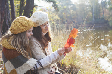 Teenager-Mädchen in Strickwaren sitzen am Fluss und betrachten lächelnd die Herbstblätter - ISF12549