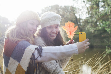 Teenager-Mädchen in Strickwaren am Fluss, die ein Herbstblatt halten und mit ihrem Smartphone ein Selfie machen und dabei lächeln - ISF12551