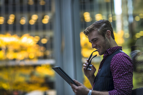 Glücklicher junger Geschäftsmann lehnt an einem Parkfenster und liest auf einem digitalen Tablet, lizenzfreies Stockfoto