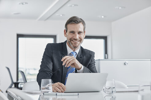 Portrait of smiling businessman with laptop at desk in office - RORF01343
