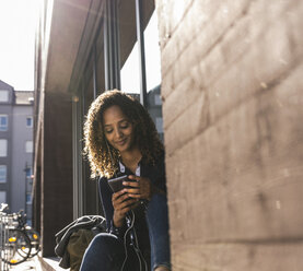 Junge Frau sitzt vor einem Fenster in der Stadt und benutzt ein Smartphone - UUF14150