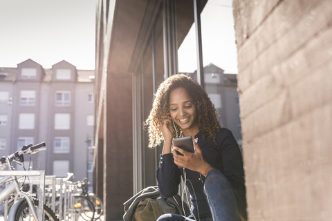 Junge Frau sitzt vor einem Fenster in der Stadt und benutzt ein Smartphone, lizenzfreies Stockfoto