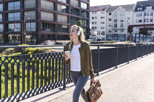 Junge Frau, die auf einer Brücke spazieren geht, Kaffee trinkt und mit Kopfhörern Musik hört - UUF14157
