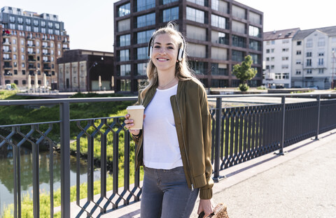Junge Frau, die auf einer Brücke spazieren geht, Kaffee trinkt und mit Kopfhörern Musik hört, lizenzfreies Stockfoto