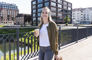 Junge Frau, die auf einer Brücke spazieren geht, Kaffee trinkt und mit Kopfhörern Musik hört - UUF14158