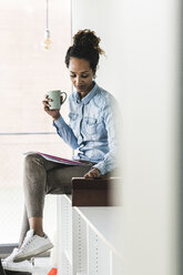 Young businesswoman sitting on shelf, using digital tablet, drinking coffee - UUF14184