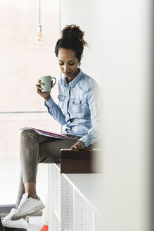 Junge Geschäftsfrau, die auf einem Regal sitzt, ein digitales Tablet benutzt und Kaffee trinkt - UUF14184