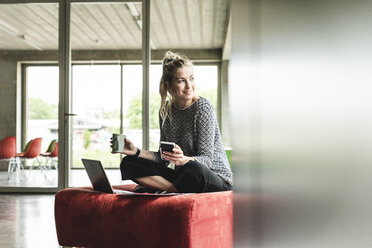 junge Geschäftsfrau arbeitet in einem modernen Büro, sitzt auf einem Hocker und benutzt einen Laptop - UUF14206