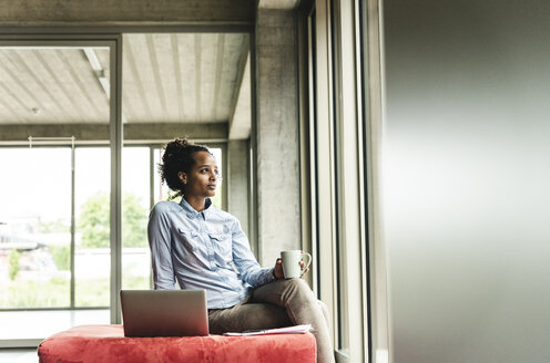 junge Geschäftsfrau arbeitet in einem modernen Büro, sitzt auf einem Hocker und benutzt einen Laptop - UUF14212