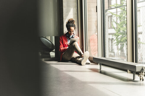 Junge Frau mit Kopfhörern, die auf dem Boden sitzt und ein digitales Tablet benutzt - UUF14217
