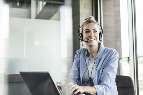 Junge Geschäftsfrau am Schreibtisch sitzend, telefonierend, mit Headset und Laptop - UUF14231