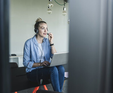 Junge Geschäftsfrau am Schreibtisch sitzend, telefonierend, mit Headset und Laptop - UUF14235